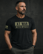 A man wearing a black graphic t-shirt with a distressed beige print that reads 'AT WAR WITH MYSELF' on the chest, standing against a dark textured wall.