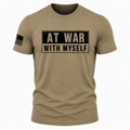A short-sleeve crewneck t-shirt in heathered tan/khaki, featuring a distressed black graphic on the chest that reads 'AT WAR WITH MYSELF' in beige block letters. A black American flag graphic is printed on the right sleeve.
