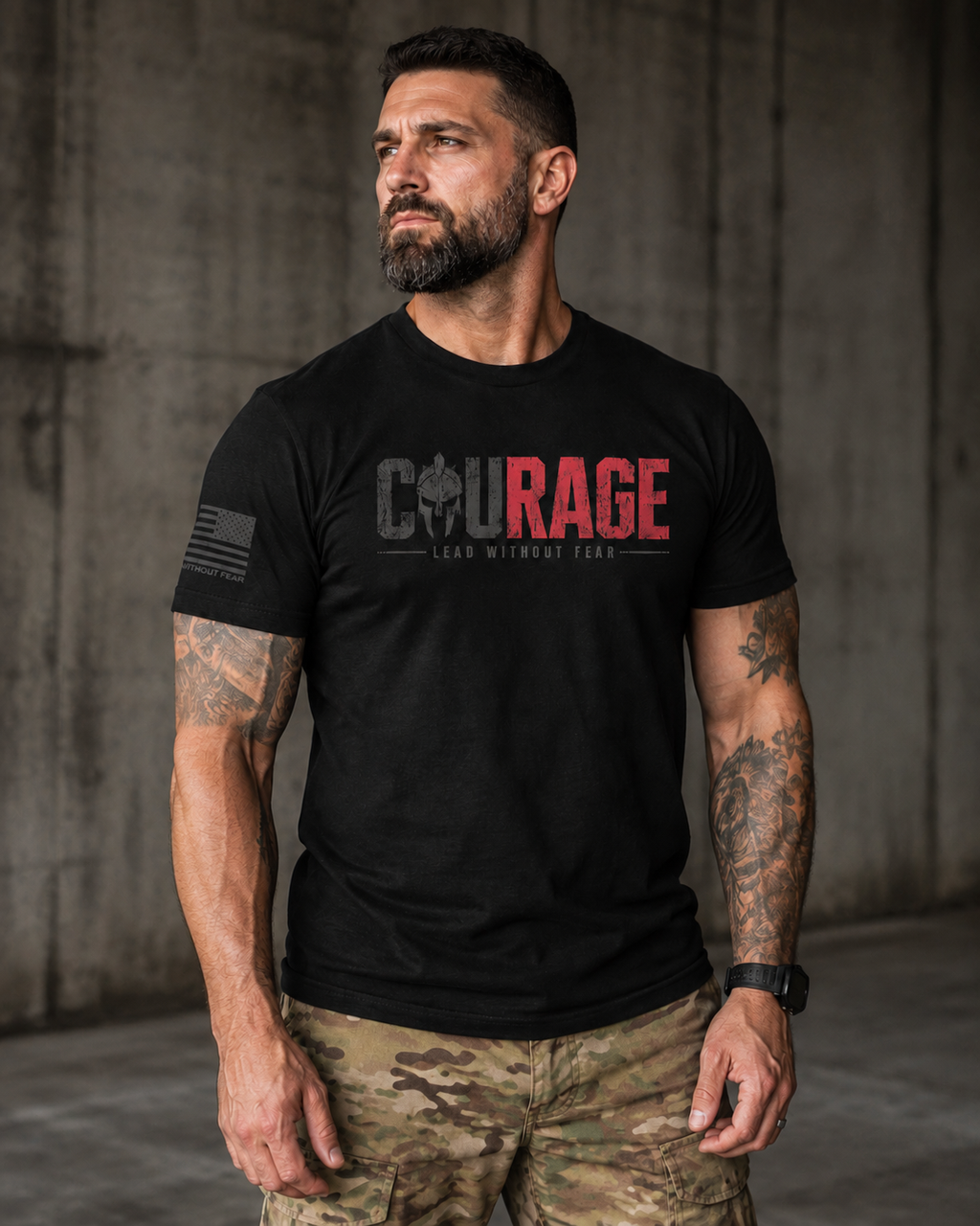 A man wearing a black short-sleeve t-shirt with a red and gray graphic on the chest that reads 'COURAGE LEAD WITHOUT FEAR' and camouflage cargo pants, standing against a concrete wall.