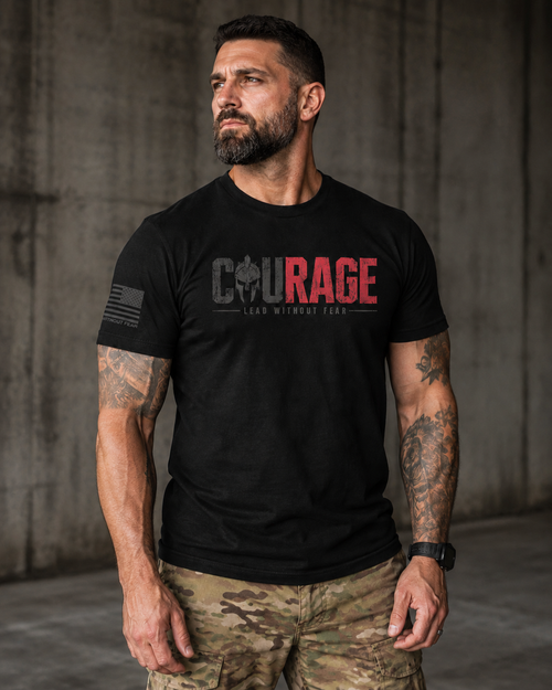 A man wearing a black short-sleeve t-shirt with a red and gray graphic on the chest that reads 'COURAGE LEAD WITHOUT FEAR' and camouflage cargo pants, standing against a concrete wall.