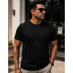 A man wearing a plain black short-sleeve crewneck t-shirt and beige pants, standing outdoors in front of a building with a glass storefront.