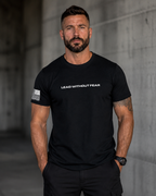 A muscular man with a beard wearing a black crewneck t-shirt with 'LEAD WITHOUT FEAR' printed in white across the chest and a distressed American flag graphic on the right sleeve, standing against a concrete wall.