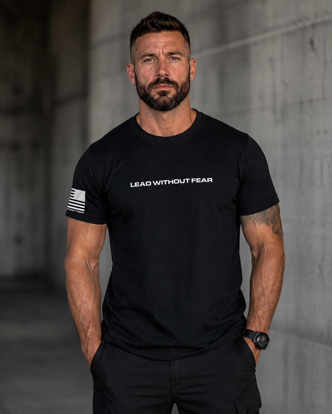 A muscular man with a beard wearing a black crewneck t-shirt with 'LEAD WITHOUT FEAR' printed in white across the chest and a distressed American flag graphic on the right sleeve, standing against a concrete wall.