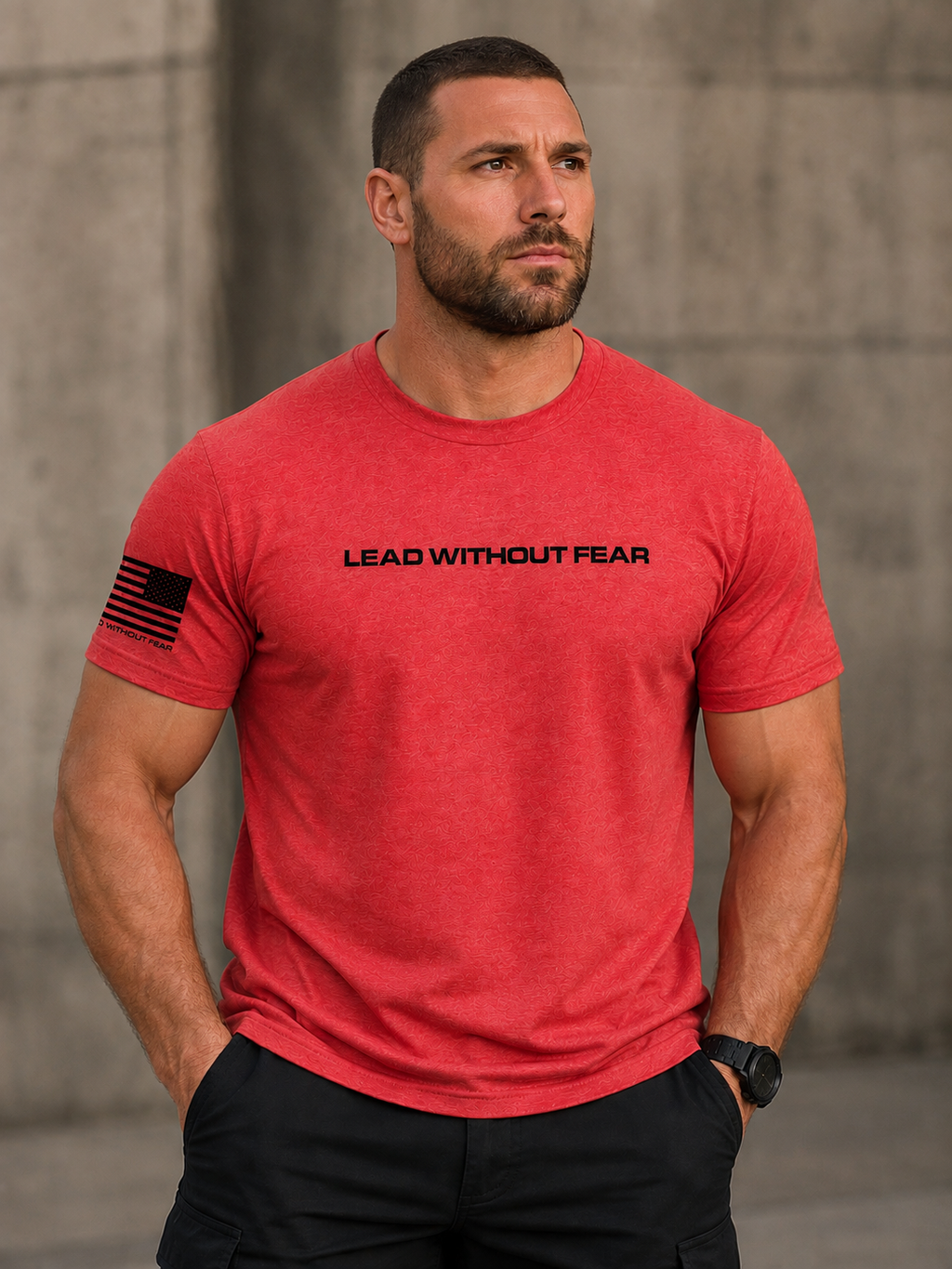 A muscular man wearing a heather red short-sleeve t-shirt with 'LEAD WITHOUT FEAR' printed in black across the chest and a black American flag graphic on the sleeve, set against a concrete wall background.