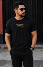 A man wearing a black short-sleeve t-shirt with a gold winged logo and text on the chest, paired with black pants, standing outdoors against a dark corrugated metal wall.