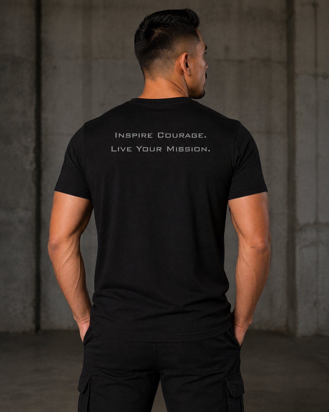 A man viewed from behind wearing a black short-sleeve t-shirt with the white printed text 'INSPIRE COURAGE, LIVE YOUR MISSION.' on the back, set against a dark concrete wall background.