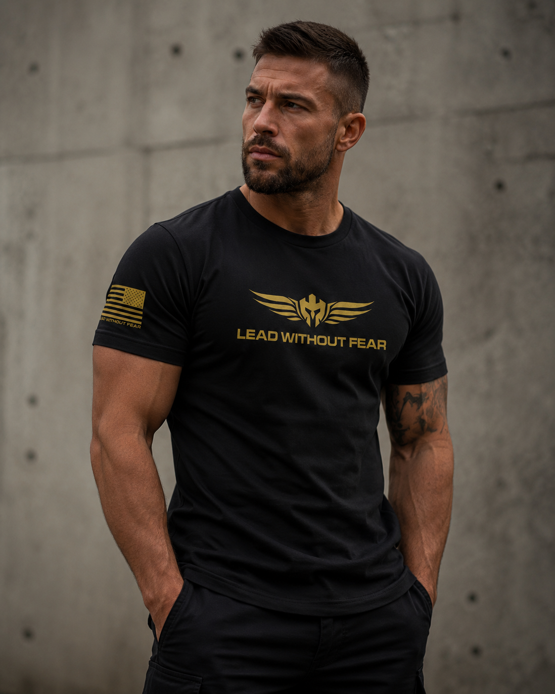A muscular man wearing a black t-shirt with a gold winged logo and the text 'LEAD WITHOUT FEAR' on the chest, standing against a concrete wall.