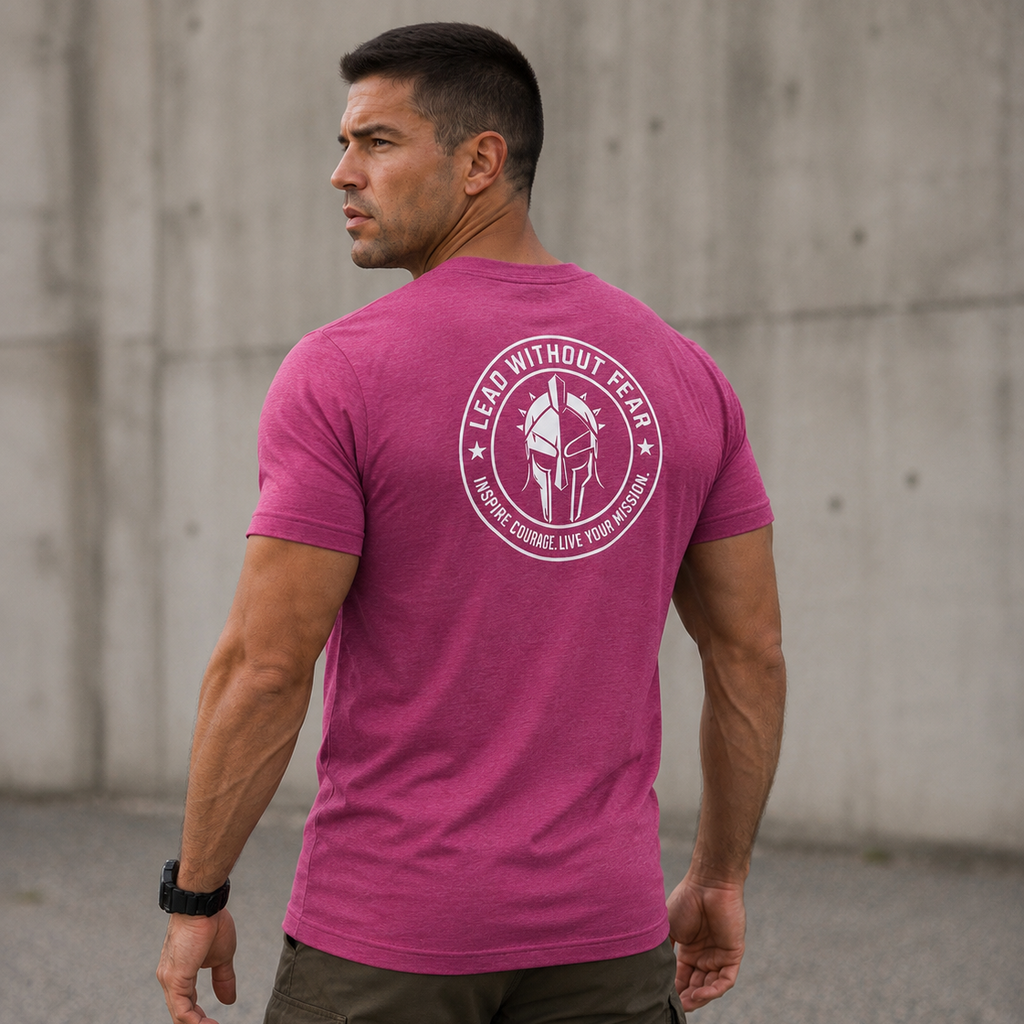 A man wearing a heather magenta short-sleeve t-shirt with a circular white graphic on the back, paired with olive cargo pants, standing in front of a concrete wall.