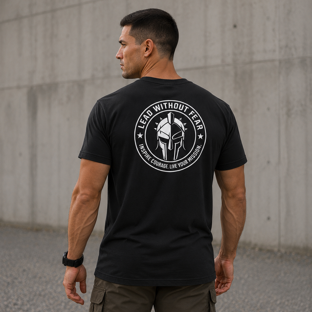 A man wearing a black t-shirt with a white circular graphic on the back, standing outdoors against a concrete wall.