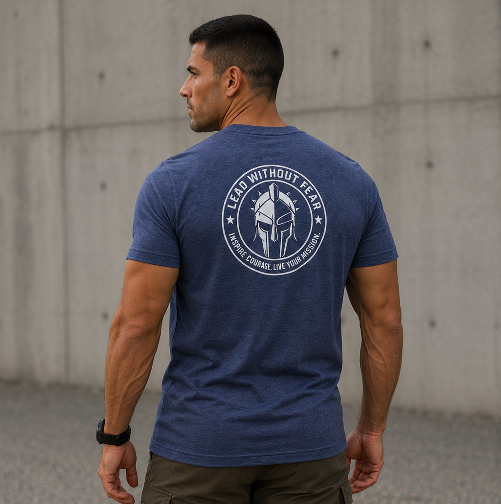 A man wearing a heathered blue t-shirt with a white circular graphic on the back, standing outdoors against a concrete wall.