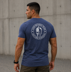 A man wearing a heathered blue t-shirt with a white circular graphic on the back, standing outdoors against a concrete wall.