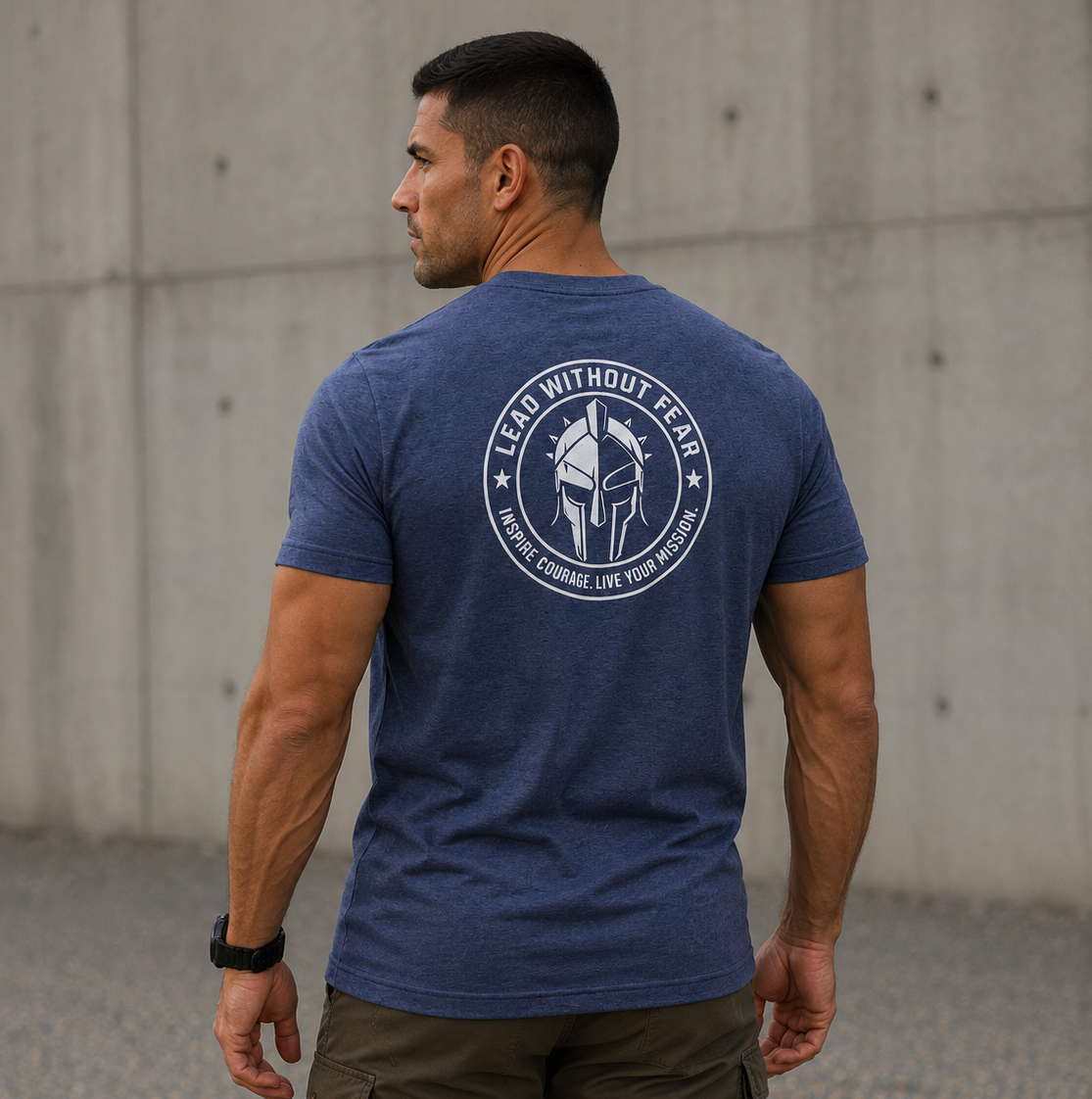 A man wearing a heathered blue t-shirt with a white circular graphic on the back, standing outdoors against a concrete wall.