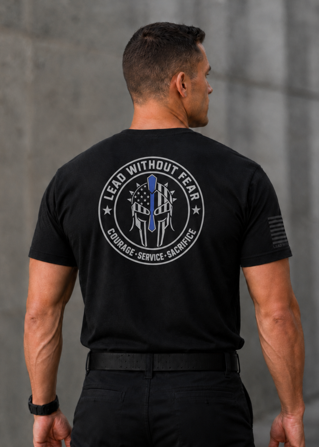 A man viewed from behind wearing a black short-sleeve tactical T-shirt with a large circular back graphic that reads 'LEAD WITHOUT FEAR' and 'COURAGE - SERVICE - SACRIFICE' around a police badge design. The shirt also has a small black flag-style print on the right sleeve.
