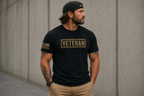 A man wearing a black t-shirt with a gold 'VETERAN' graphic on the chest and a black baseball cap, standing against a concrete wall.