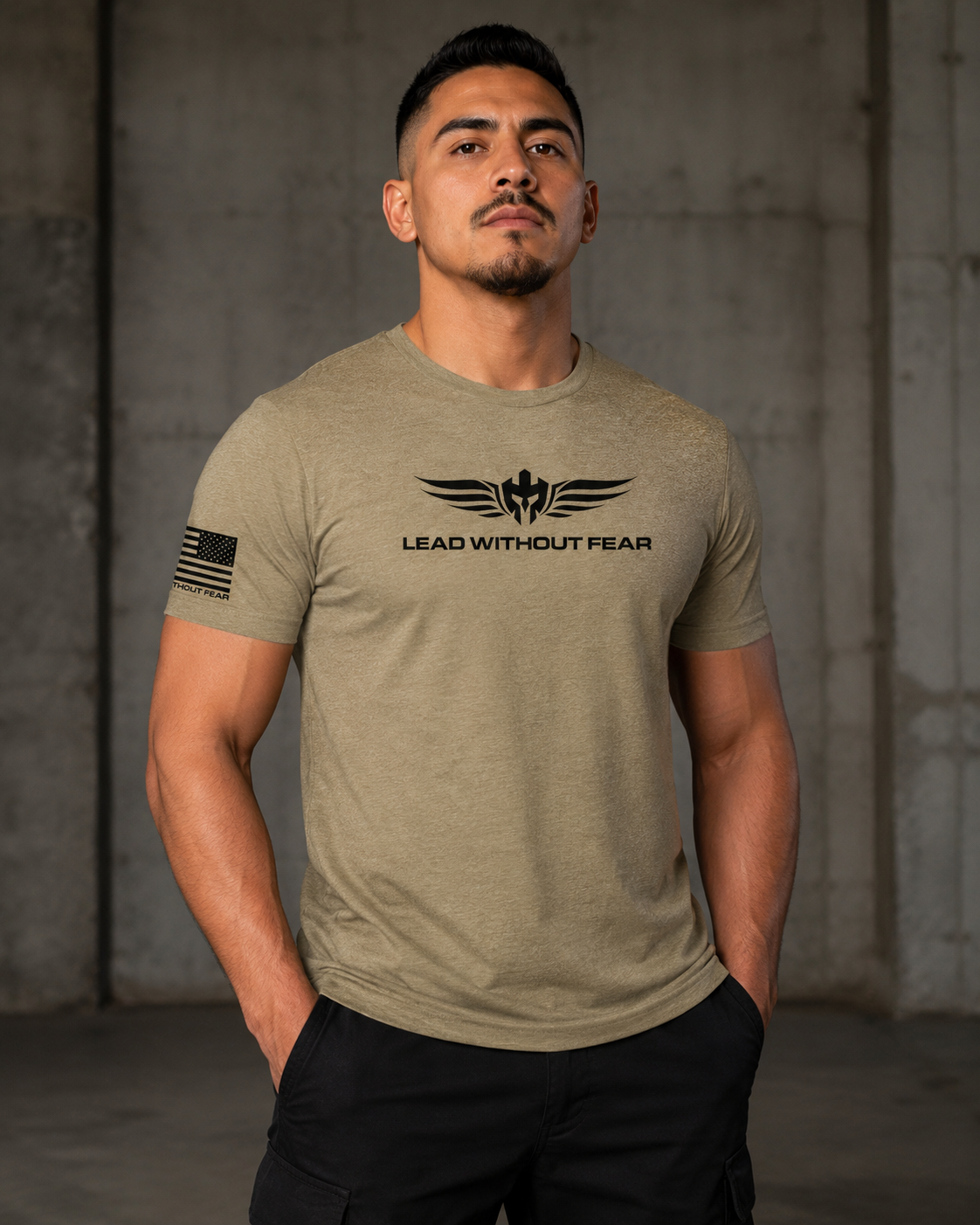 A man wearing a heathered khaki t-shirt with a black winged logo and the text 'LEAD WITHOUT FEAR' on the chest, along with a black American flag graphic on the sleeve, standing against a concrete wall.