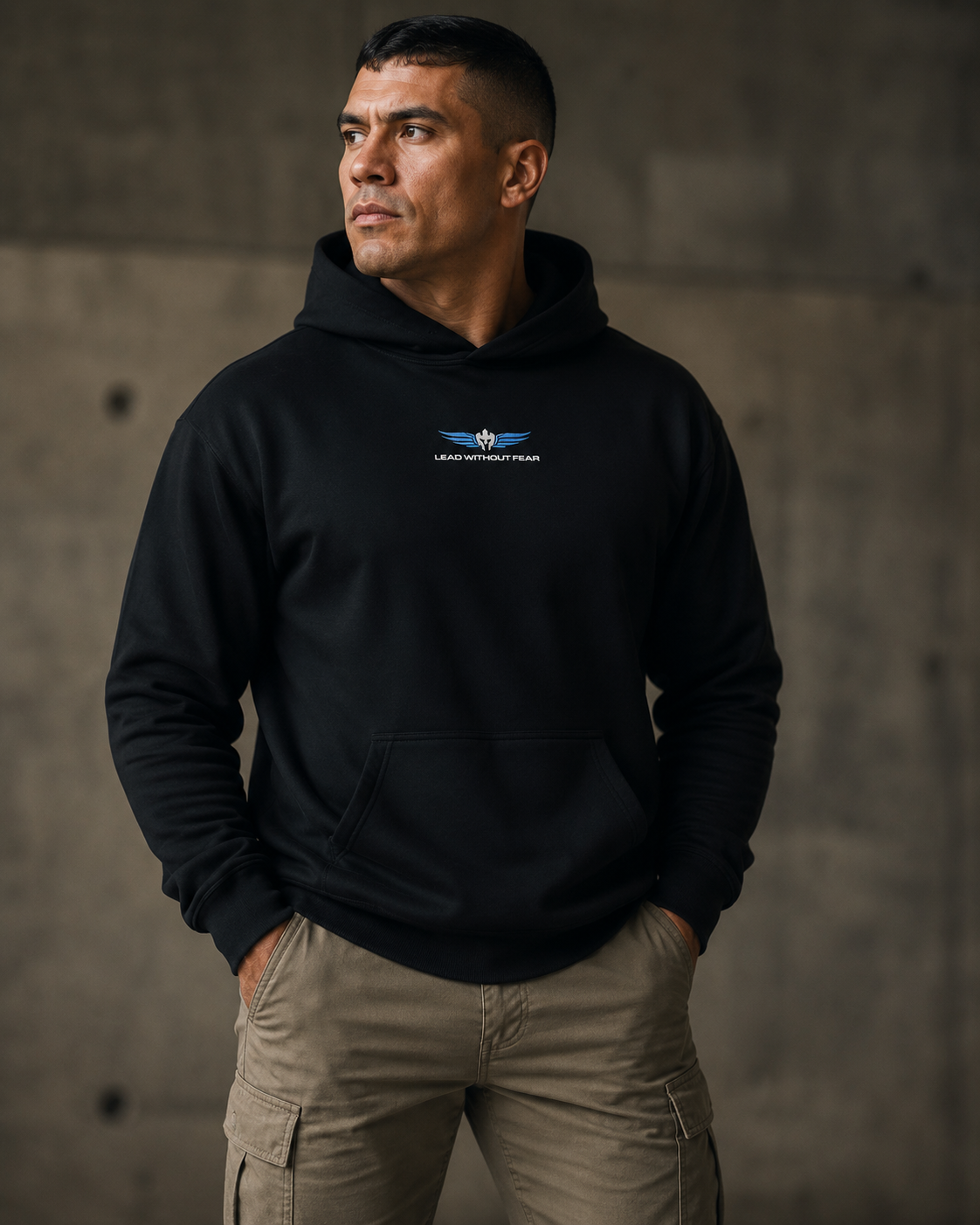 A man wearing a black pullover hoodie with a small Lead Without Fear logo on the center chest, paired with khaki cargo pants, standing against a textured concrete wall.