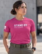 A woman wearing a bright pink t-shirt with 'STAND BY' printed in white across the chest, paired with olive green cargo pants, standing outdoors against a blurred concrete wall.