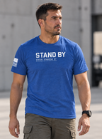 A man wearing a heather blue t-shirt with 'STAND BY' printed in white across the chest, paired with olive cargo pants, standing in an outdoor concrete setting.