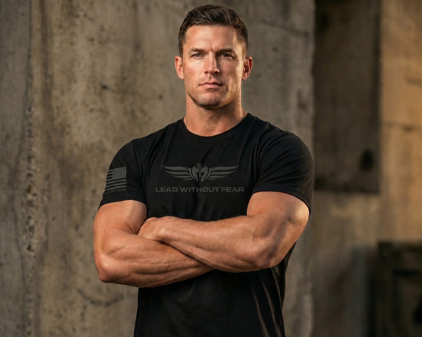 A muscular man wearing a black short-sleeve t-shirt with a gray winged logo and the text 'LEAD WITHOUT HEAR' on the chest, standing with arms crossed against a textured concrete wall.
