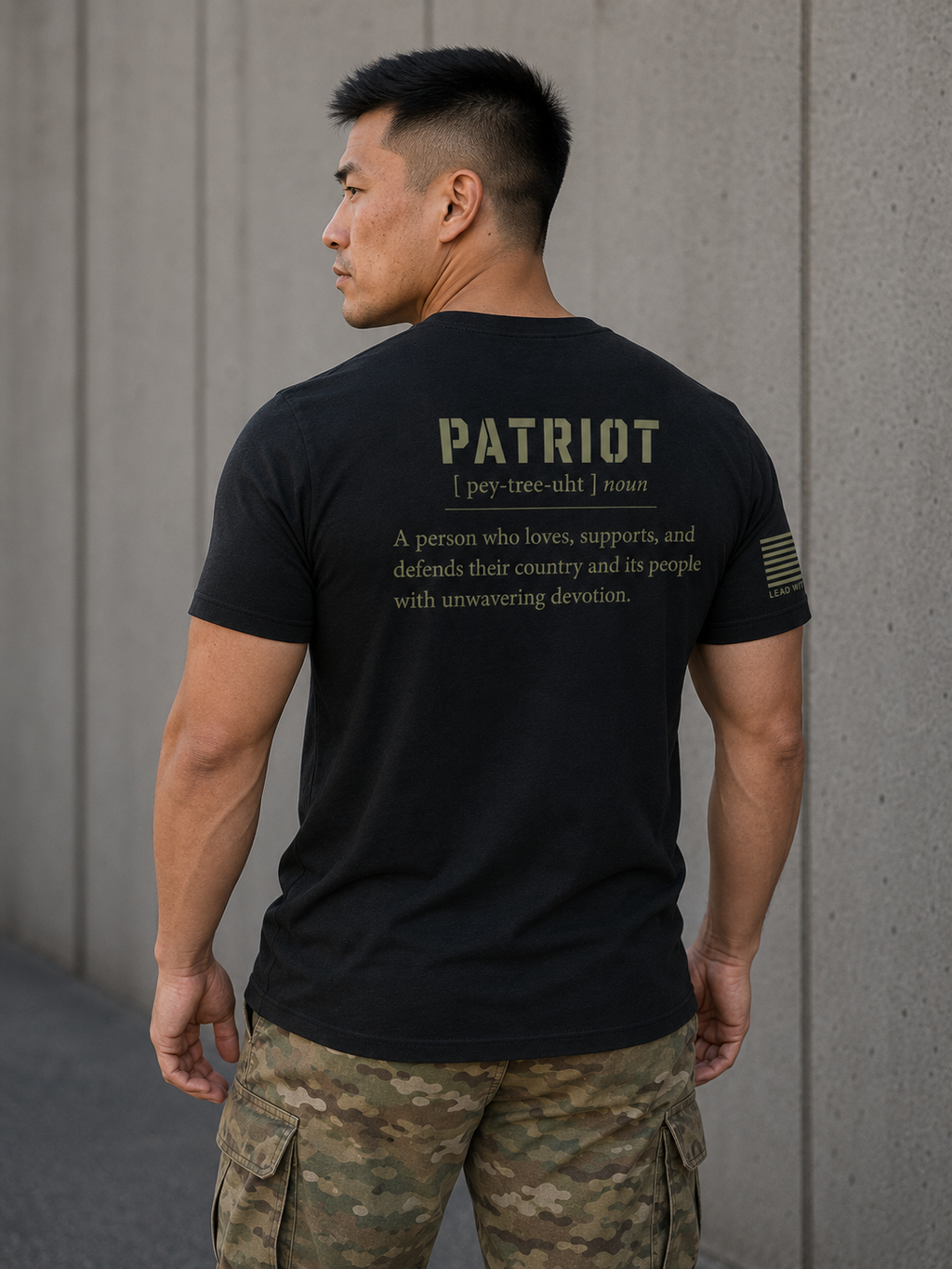 A man wearing a black t-shirt with a military-style 'PATRIOT' dictionary definition printed on the back, paired with camouflage cargo pants, standing against a concrete wall.