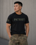 A man wearing a black short-sleeve t-shirt with 'PATRIOT' printed in olive green across the chest and a small American flag patch on the sleeve, paired with camouflage cargo pants, standing against a concrete wall.