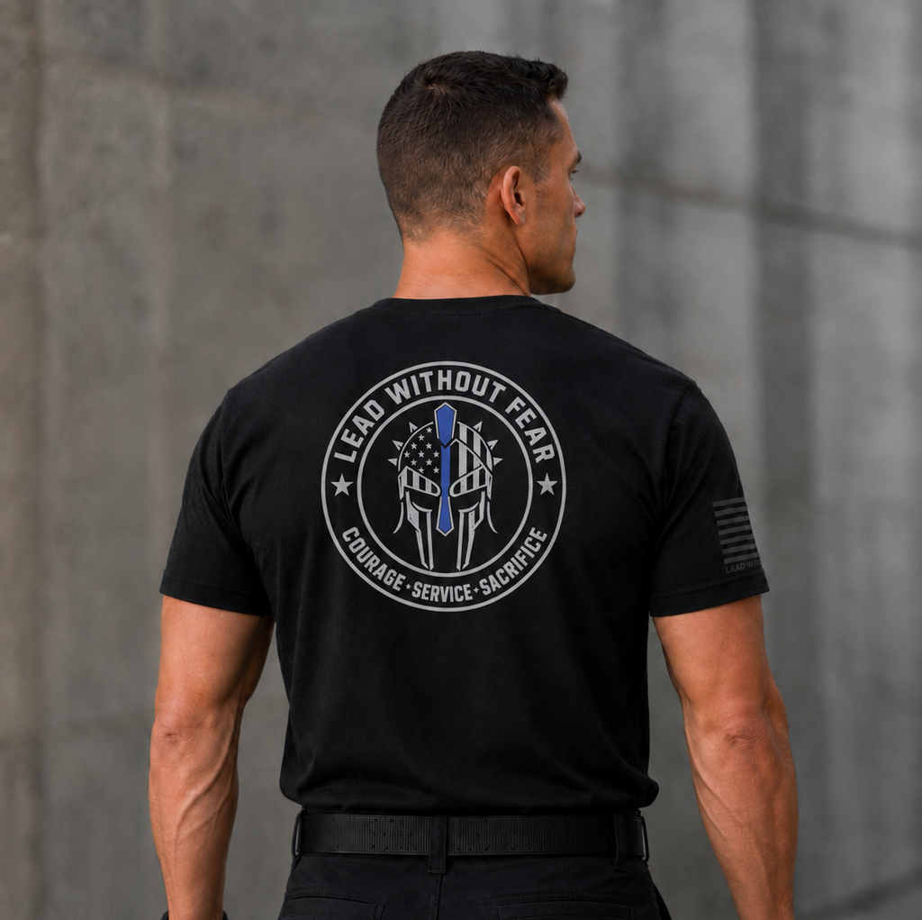 A man viewed from behind wearing a black short-sleeve tactical T-shirt with a large circular back graphic that reads 'LEAD WITHOUT FEAR' and 'COURAGE - SERVICE - SACRIFICE' around a police badge design. The shirt also has a small black flag-style print on the right sleeve.