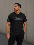 A man wearing a black short-sleeve t-shirt with a winged emblem with thin blue line on feathers and the text 'LEAD WITHOUT FEAR' on the chest, standing against a gray concrete wall.