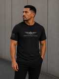 A man wearing a black short-sleeve t-shirt with a winged emblem with thin green line on feathers and the text 'LEAD WITHOUT FEAR' on the chest, standing against a gray concrete wall.
