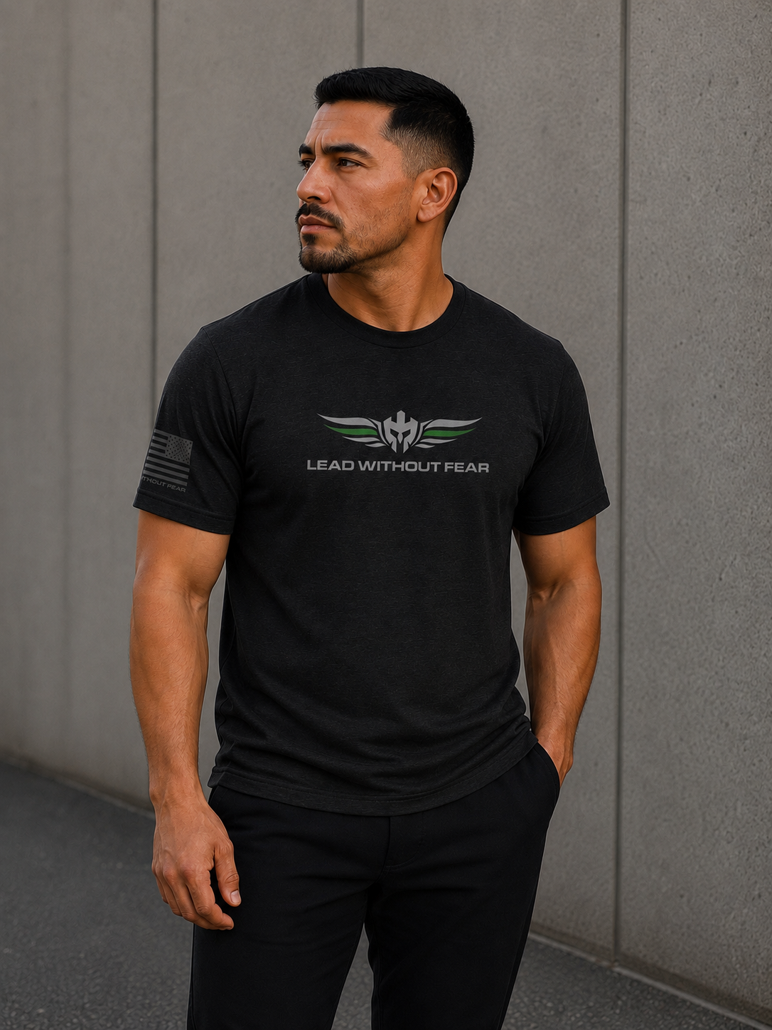 A man wearing a black short-sleeve t-shirt with a winged emblem with thin green line on feathers and the text 'LEAD WITHOUT FEAR' on the chest, standing against a gray concrete wall.
