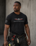 A man wearing a black short-sleeve t-shirt with a winged emblem with thin red line on feathers and the text 'LEAD WITHOUT FEAR' on the chest, standing against a gray concrete wall.