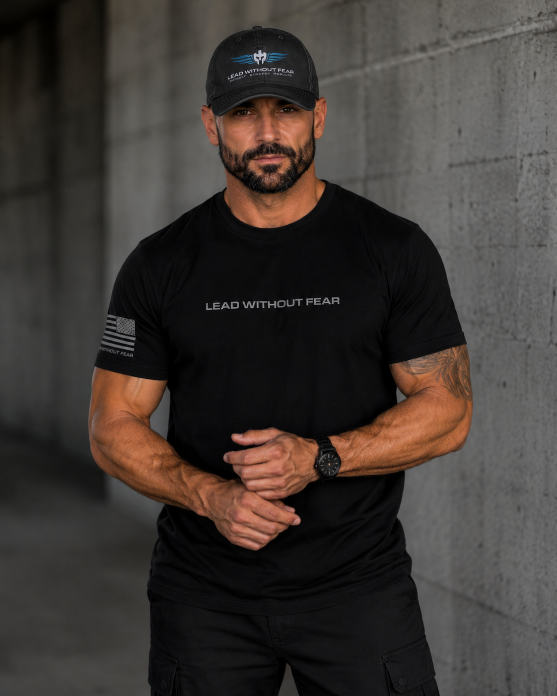 A muscular man wearing a black t-shirt with 'LEAD WITHOUT FEAR' printed on the chest, a black cap with a logo, and black cargo pants, standing against a gray concrete wall.