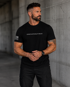 A muscular man wearing a black short-sleeve t-shirt with 'LEAD WITHOUT FEAR' printed in white across the chest and a distressed American flag graphic on the sleeve, standing against a concrete wall.