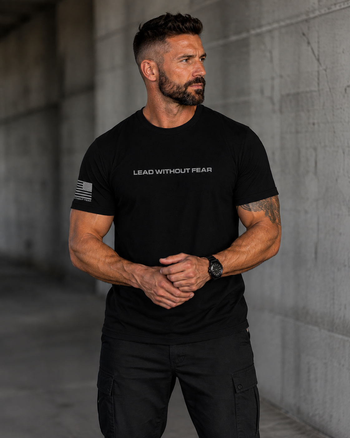 A muscular man wearing a black short-sleeve t-shirt with 'LEAD WITHOUT FEAR' printed in white across the chest and a distressed American flag graphic on the sleeve, standing against a concrete wall.