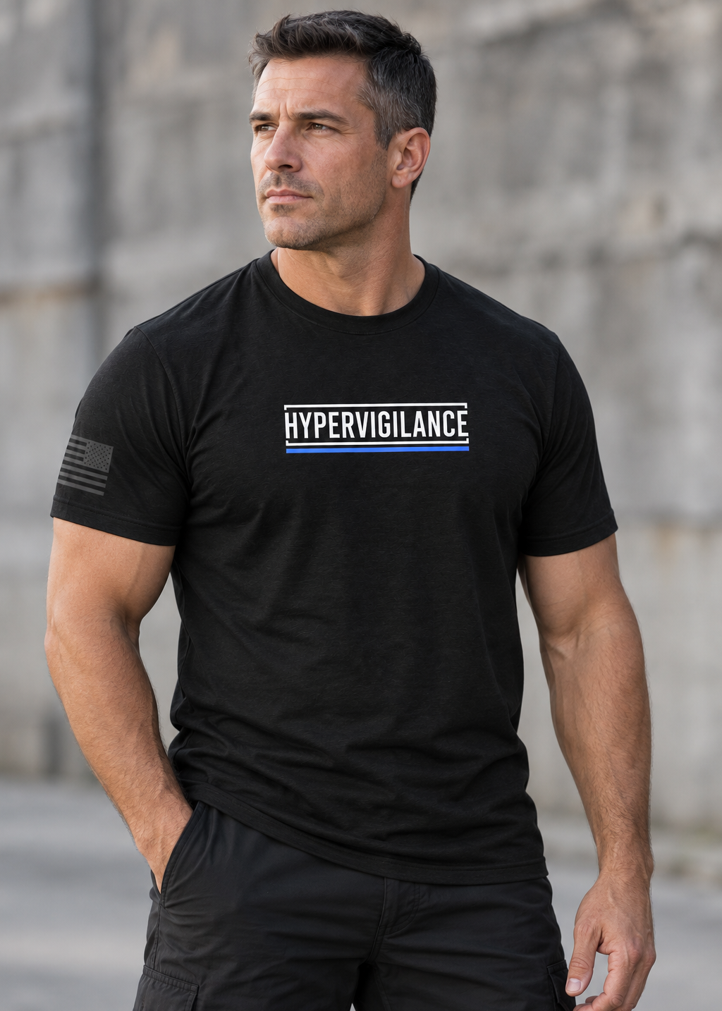 A muscular man wearing a black short-sleeve t-shirt with 'HYPERVIGILANCE' printed in white across the chest, flanked by two blue stripes, and a black flag graphic on the sleeve. He is standing outdoors against a blurred stone wall.
